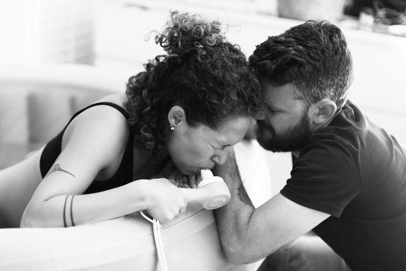 Picture of labour person in birth pool breathing in Nitrous Oxide from a mouth piece. Their partner is leaning in to them and their heads are touching.